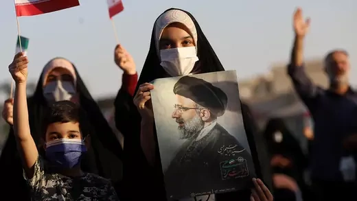 A young Iranian woman holds a portrait of Ebrahim Raisi. Other adults and a child are seen, some with small Iranian flags.