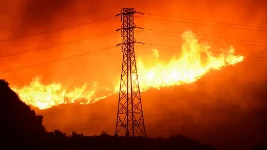 A wind-driven wildfire burns near power line tower in Sylmar, California. Flames surround the background of the electricity grid.