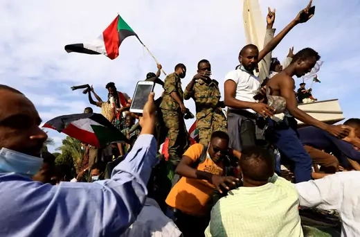 Civilians, some with Sudanese flags, climb on top of a military vehicle. Some members of the military wearing camouflage are intermixed with the protesters.