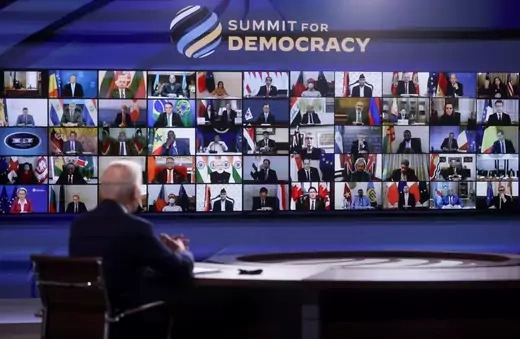 U.S. President Joe Biden sits before a panel of screens with leaders of various countries. A stylized logo of a globe next to "Summit for Democracy" is above the panel.