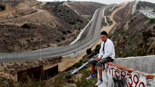 A young man, wearing a white jacket, shorts, sneakers, and a hat, sits atop a wall next to the U.S.-Mexico border wall. 