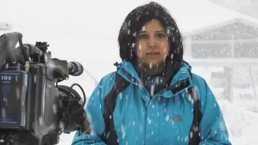 Bahar Dutt stands behind a TV camera as it snows. 