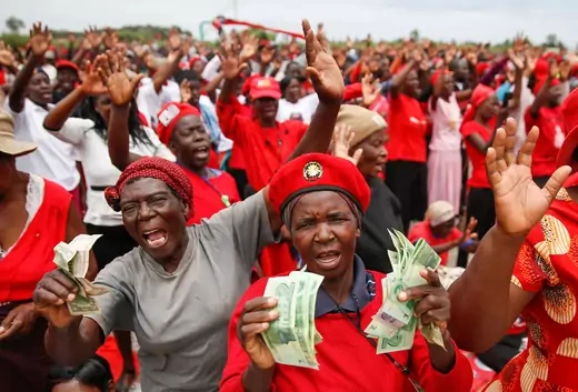 A crowd of people, mostly wearing the color red, at a rally. Two women at the front of the picture hold money in their hands for the camera.