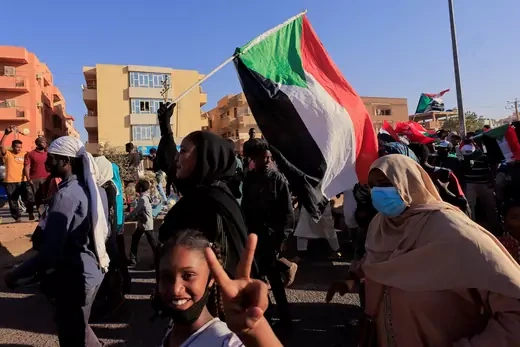 Protesters, one holding a Sudanese flag, walk down the street. A young girl in the foreground throws up two fingers in a peace symbol.