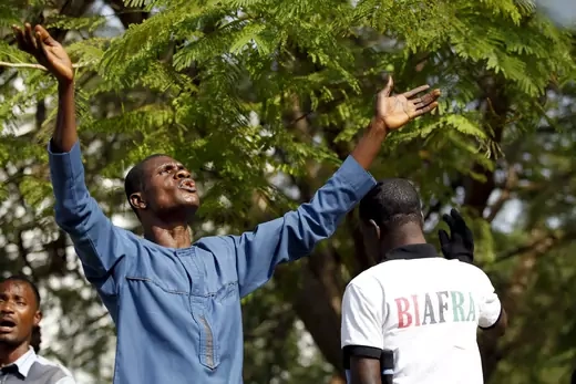 A man looks up at the sky with his arms outstretched. Another man walks by him, wearing a shirt with "Biafra" written across the back.