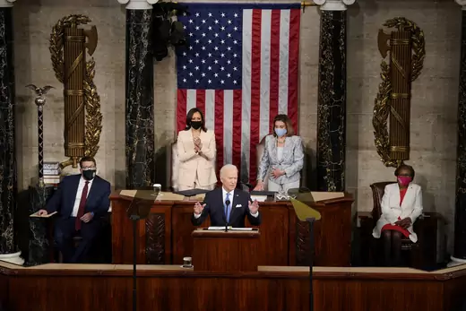 President Joe Biden gestures while speaking in the House Chamber. He is flanked by two House staff members, Vice President Kamala Harris, and Speaker Nancy Pelosi.