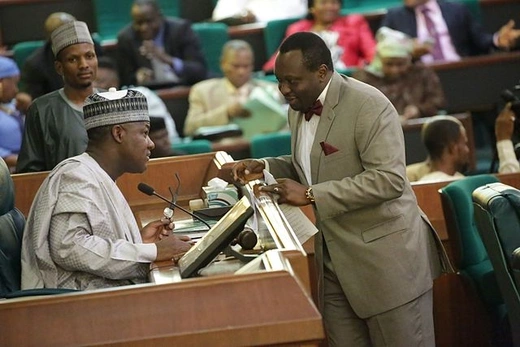 A member of the Nigerian House of Representatives wearing a brown suit speaks to a man sitting behind a desk wearing a traditional Nigerian outfit. 