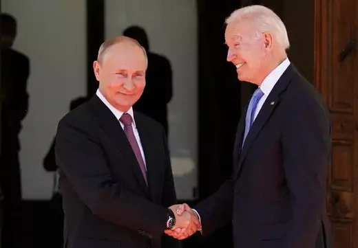 U.S. President Joe Biden and Russian President Vladimir Putin shake hands in front of a glass door.