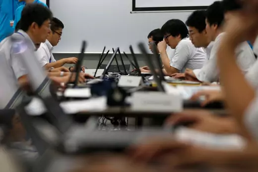 A group of cybersecurity personnel sit facing each other at a row of laptops.