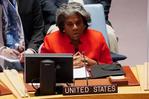 United States Ambassador to the United Nations wearing a red jacket speaks into a microphone while seated at the United Nations. 