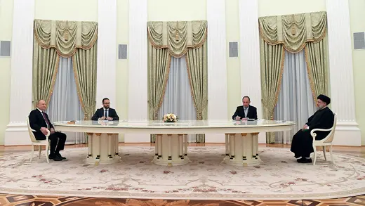 Vladimir Putin and Ebrahim Raisi sit on two ends of a long, white table. Two men sit between them.