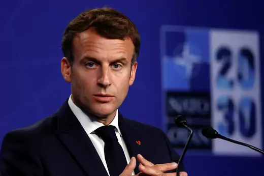 French President Emmanuel Macron stands at a podium with his hands folded.