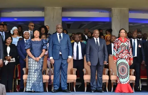 The incoming and outgoing Congolese presidents stand wearing official dress next to their wives. A group of officials and other individuals stand behind them.