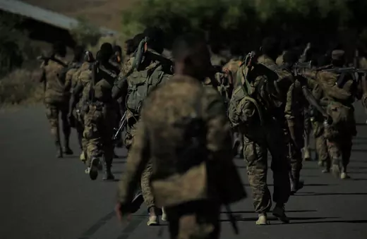 Military troops wearing camouflage are seen from behind walking in a large group.