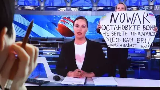 A woman views a computer screen that shows a woman holding a "No War" poster and standing behind an anchorwoman at a news desk