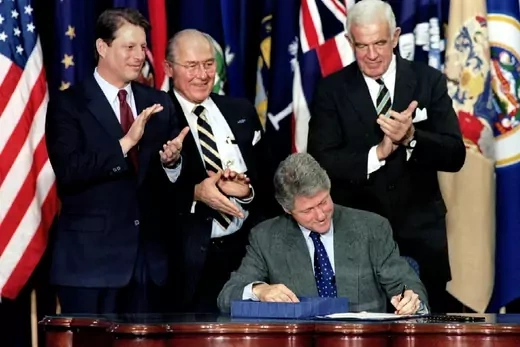 U.S. President Clinton sitting at desk surrounded by politicians, signs the NAFTA agreement.