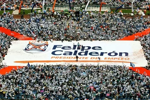 Large banner with writing saying "Felipe Calderon" is held up by crowd of supporters.