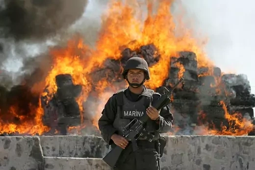 A Mexican marine holding a machine gun stands in front of flames from a burning pile of cocaine