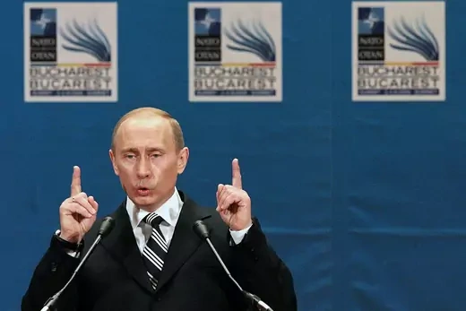 Russian President Vladimir Putin holds up both index fingers while speaking into microphone.