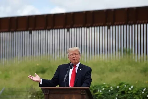 President Trump stands in front of unfinished border walk, talking into a microphone.