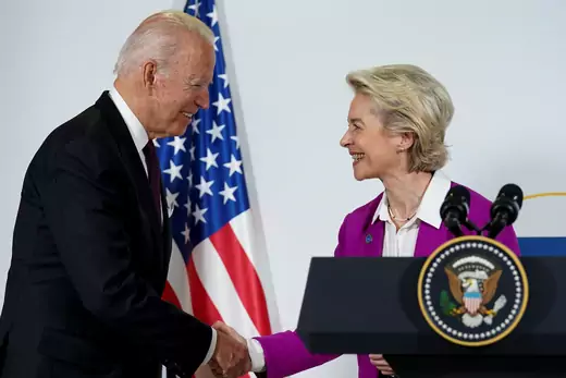 President Joe Biden and European Commission President Ursula von der Leyen shake hands at a podium in front of an American flag.