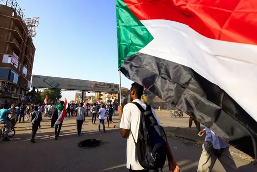 Man in white shirt carries large Sudanese flag during protests in the capital city, Khartoum.