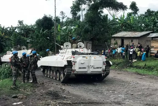 Soldiers wearing camouflage and blue helmets are seeing in front of a white tank. In the background, civilians gather in front of a small house.   