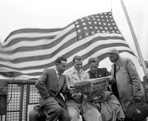4 male refugees from Hungary read a newspaper with the headline "Hungarians Battle Reds", under a big U.S. flag.