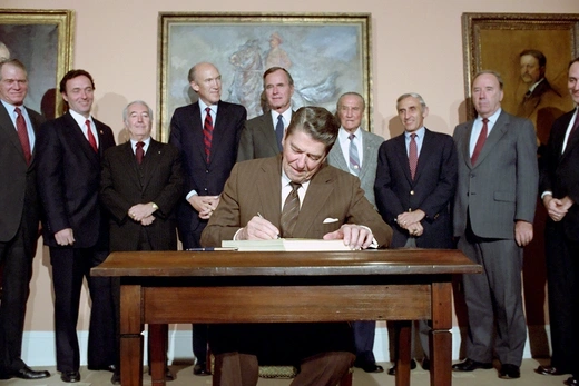 President Ronald Reagan signs the Immigration Reform Act, sitting at a desk. Standing behind him, looking on, are Dan Lungren, Strom Thurmond, George Bush, Romano Mazzoli and Alan Simpson.