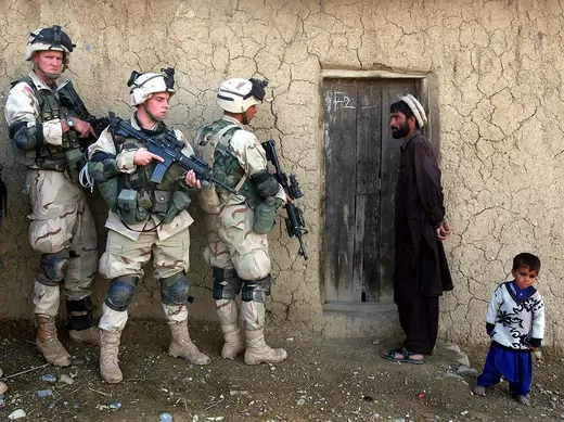 An Afghan man and his son standing in front of their wooden entrance door watch as soldiers from the U.S. Army 82nd Airborne Division prepare to enter.