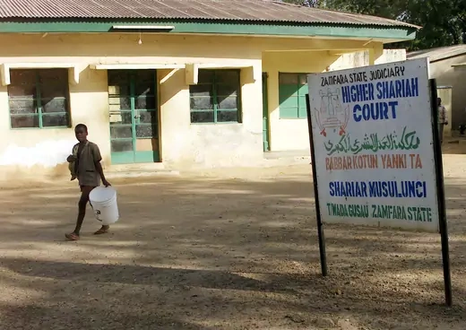 A sign that reads "Higher Shariah Court," a child with a bucket in the background, and an old looking building. 