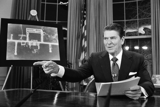 Photo of President Ronald Reagan in the Oval Office, addressing the nation while  pointing at a picture of Soviet Migs in western Cuba.