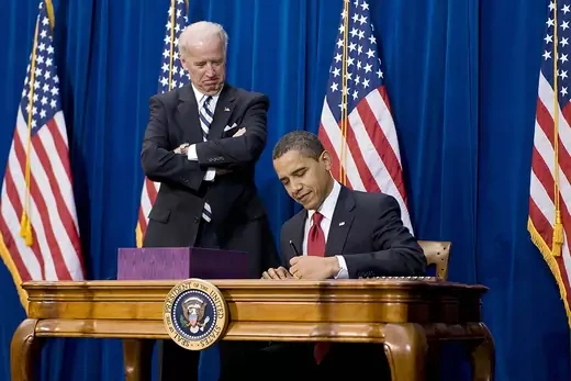 President Barack Obama sitting at desk, alongside Vice President Joseph Biden standing next to him,, signs the American Recovery and Reinvestment Act.
