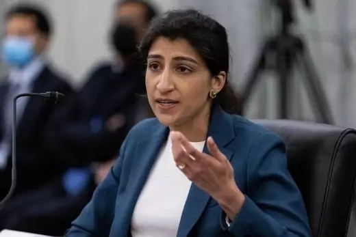 Lina Khan gestures in front of a microphone with several people in the background who are out of focus.