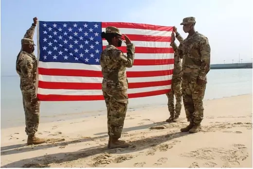 Two soldiers hold a U.S. flag behind a soldier administering an oath to another solider on a beach. 