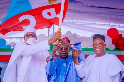 Nigeria APC presidential candidate Tinubu holds a flag up with the help of two individuals while smiling.