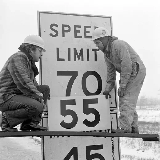 Two highway workers change a speed limit sign from 70 mph to the new federally mandated limit of 55 mph.
