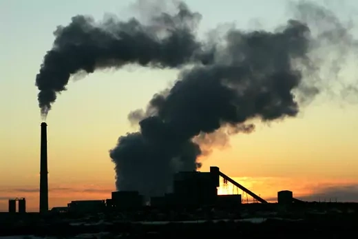 Black smoke is seen billowing from factory chimney at sunset.