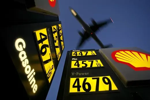 A low angle view of gas prices signs with a plane flying overhead.