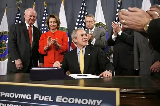 U.S. President George W. Bush is seen sitting at desk, smiling, surrounded by members of Congress, ready to sign the Energy Independence and Security Act. 
