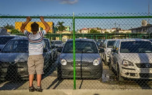 A boy looks through a fence at used cars for sale in Havana.