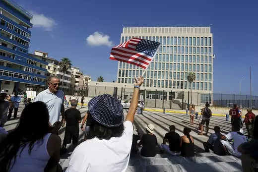 A woman facing away from the camera holds a U.S. flag above her head with the U.S. embassy in Havana in the background.