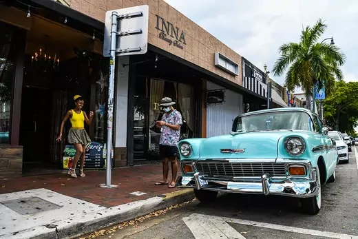Two people stand outside a restaurant next to an old old car, in a Cuban neighborhood of Miami, Florida.