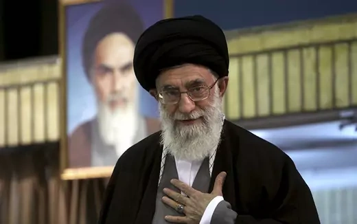 Ali Khamenei stands in front of a portrait over himself with his hand over his lapel.