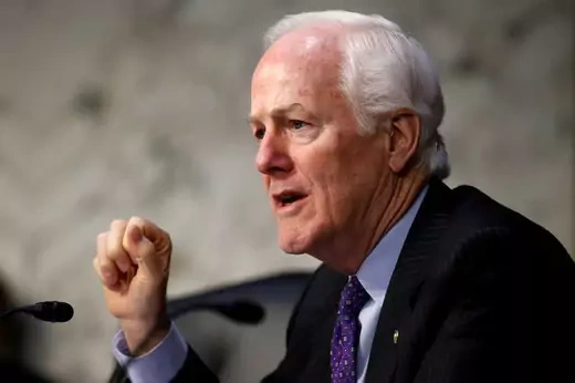 John Cornyn gestures in front of a microphone with a muddle gray background
