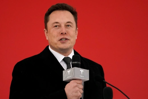 Elon Musk stands in front of an all red background while holding a microphone with the tesla logo emblazoned on it.