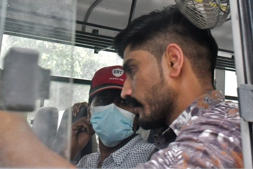 A man in a floral shirt is viewed through a partly open plexiglass window, while another man in a blue surgical mask and red hat sits next to him.