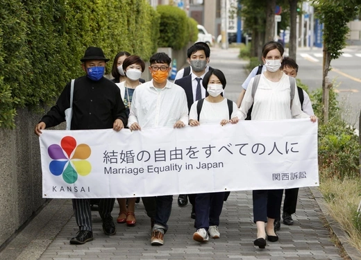 The four plaintiffs, two men and two women, walk down a sidewalk holding a banner calling for marriage equality in Japan, followed by a small group of supporters.
