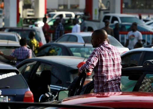 A man is outside of his car looking at a long line of vehicles in line for petrol at a gas station in Nigeria. 