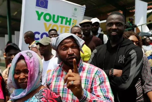 A man in a voting registration crowd in Nigeria points at the camera and is pictured in front of a youth vote poster.